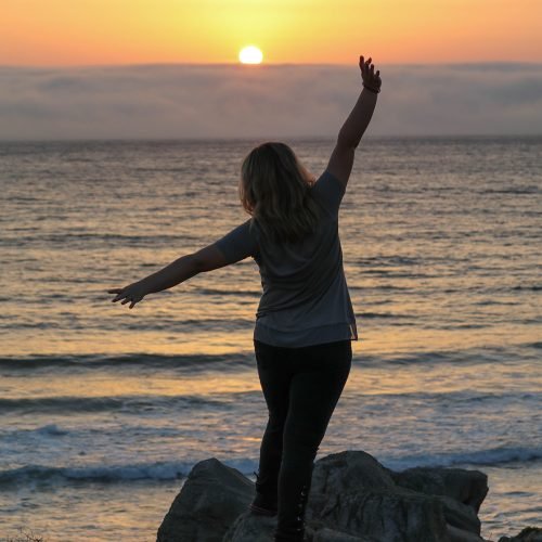 rear-view-woman-standing-rocks-with-arms-outstretched-beach